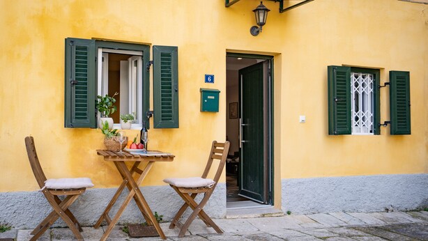 A yellow building with a wooden table and two chairs, a door, and a window with shutters. - Captivating 1-bed Apartment in Kerkira (Corfu)
