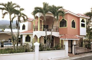 A two-story house with a balcony, surrounded by a white fence and palm trees. - Peaceful culdesac orchid villa, pool, work ergo (Puerto Plata)