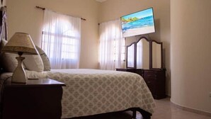 A bedroom with a bed, a nightstand, a lamp, a dresser, and a TV displaying a beach scene. - Peaceful culdesac orchid villa, pool, work ergo (Puerto Plata)