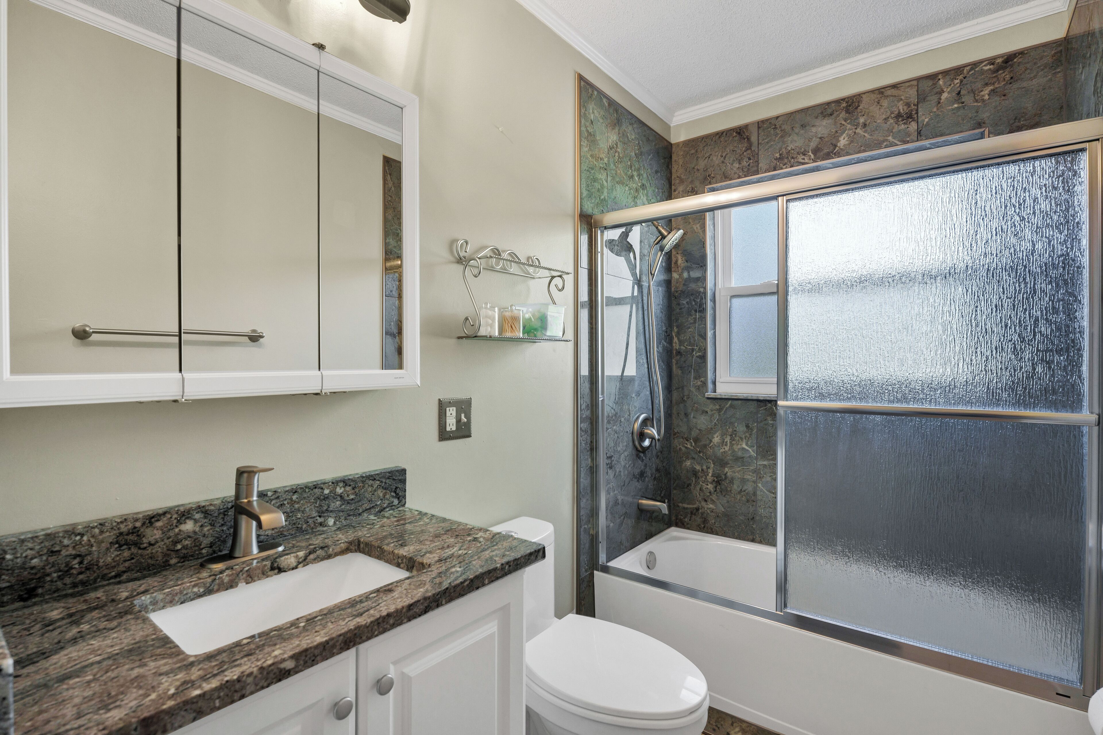 Une salle de bain avec un comptoir en granit, des toilettes blanches et une douche fermée par des parois de verre.