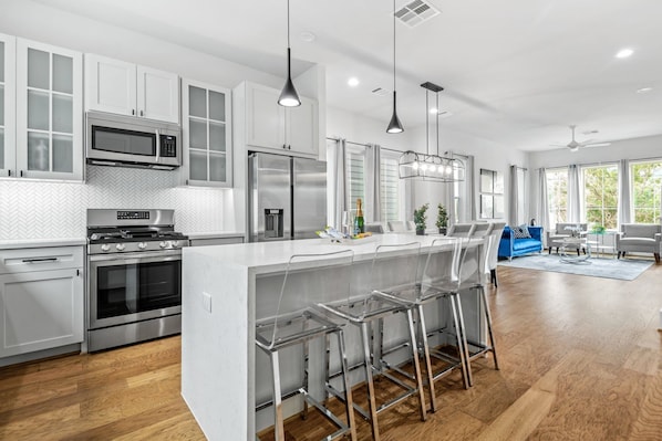 A modern kitchen with stainless steel appliances, a central island with bar stools, and a dining area with a blue sofa. - Lux Living / Rooftop / Private Chef & King Suite Downtown Oasis (Houston)
