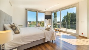 A bedroom with a large bed, a TV, and a balcony with a view of trees and buildings.
