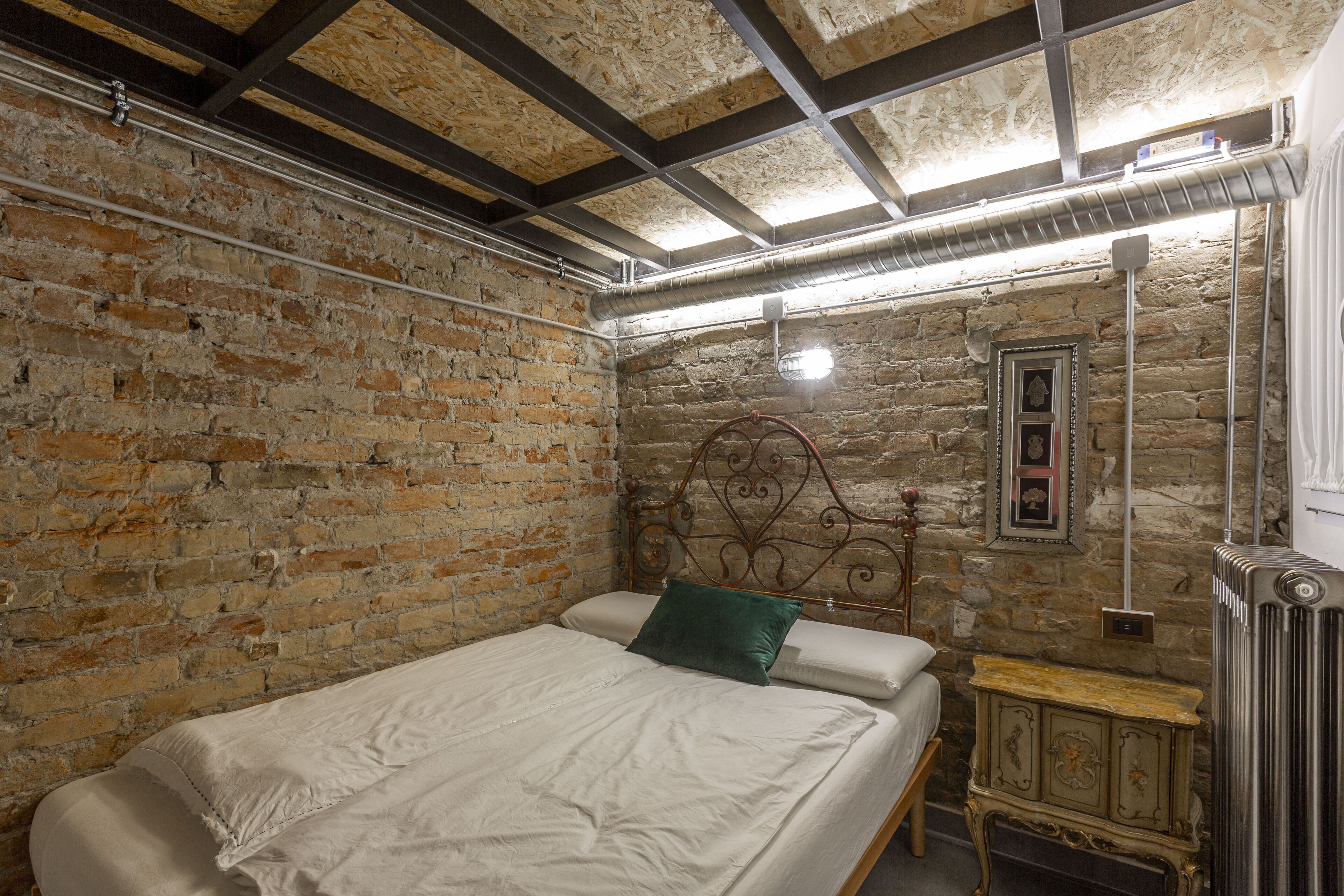 A bedroom with a brick wall, a metal headboard, a wooden nightstand, and a radiator.