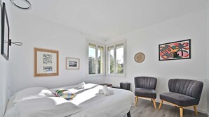 Un dormitorio con una cama, dos sillas, un ventanal y obras de arte en las paredes.