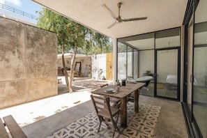 A modern patio with a wooden table and chairs, a ceiling fan, and a glass door leading to a bedroom.