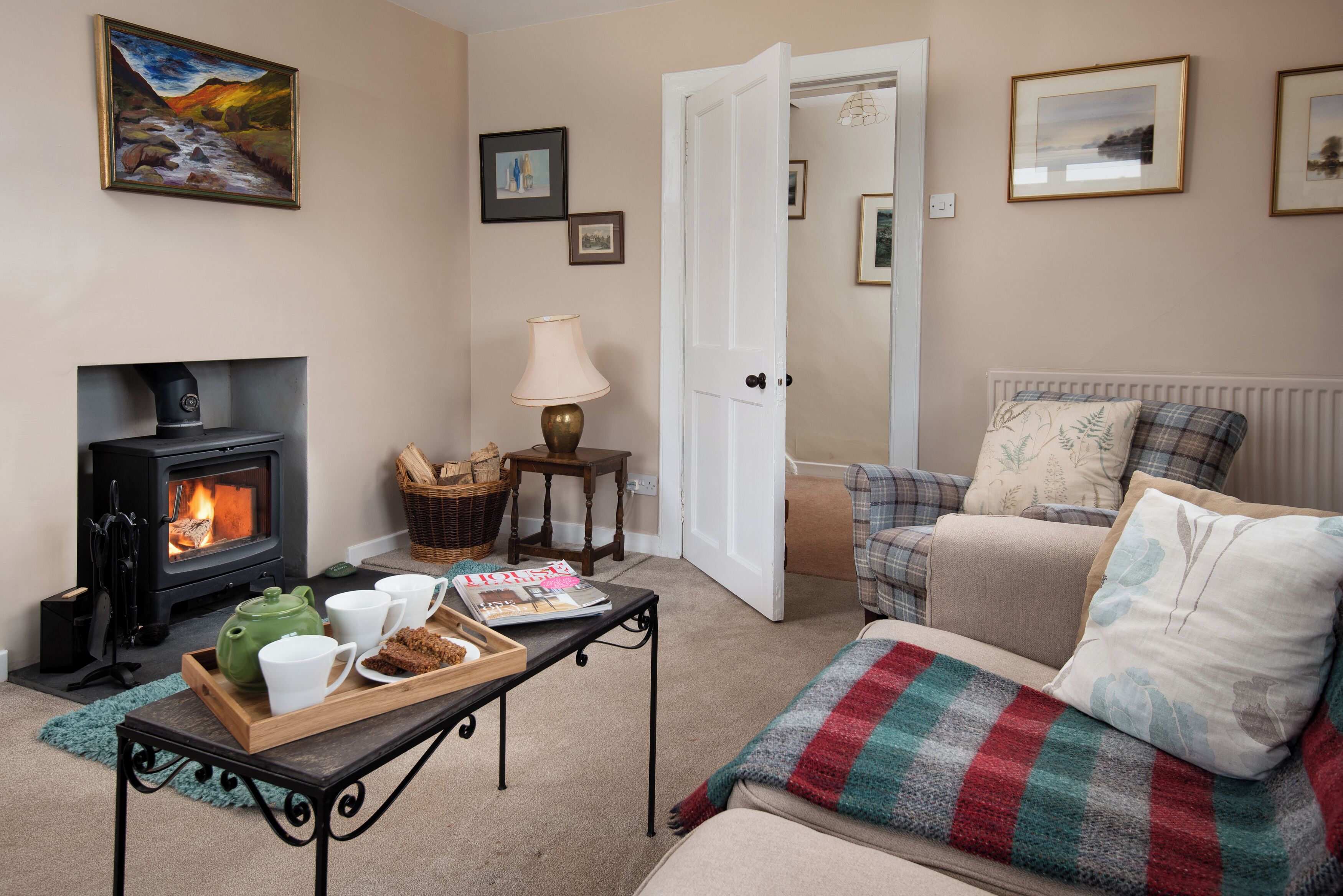 A cozy living room with a fireplace, a sofa, a coffee table with a tray of mugs, and a door leading to another room.