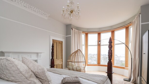 Une chambre à coucher avec un grand lit, un lustre, une vue sur la ville grâce à de grandes fenêtres et une porte en bois.