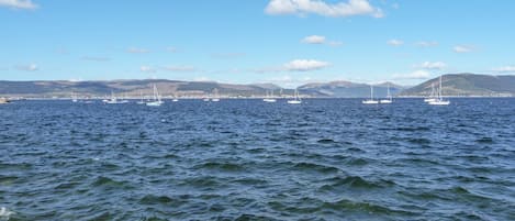 A coastal view with multiple sailboats on the water, a clear sky, and distant hills.