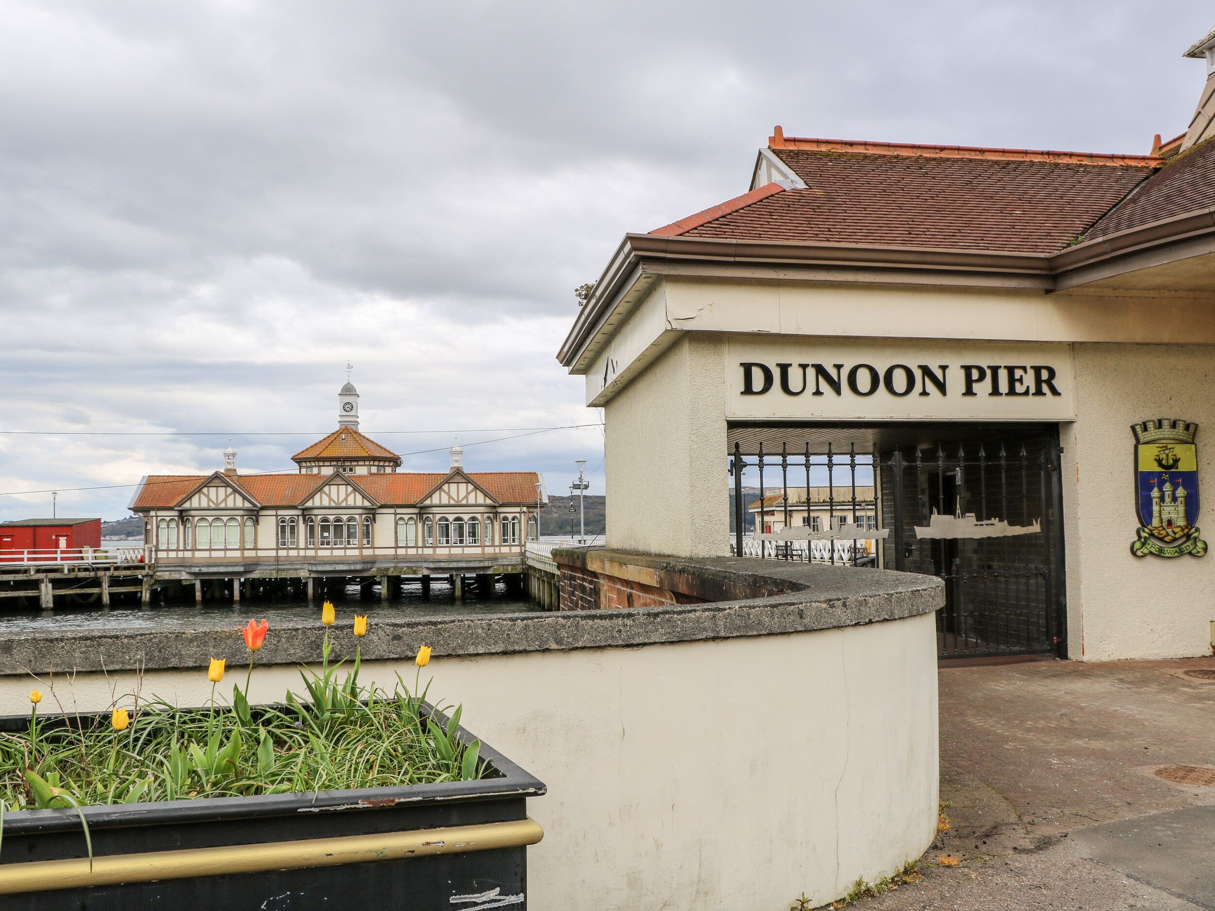 Um edifício com a placa "DUNOON PIER", estilo arquitetônico tradicional, um cais que se estende para a água e uma pequena área de jardim com tulipas.