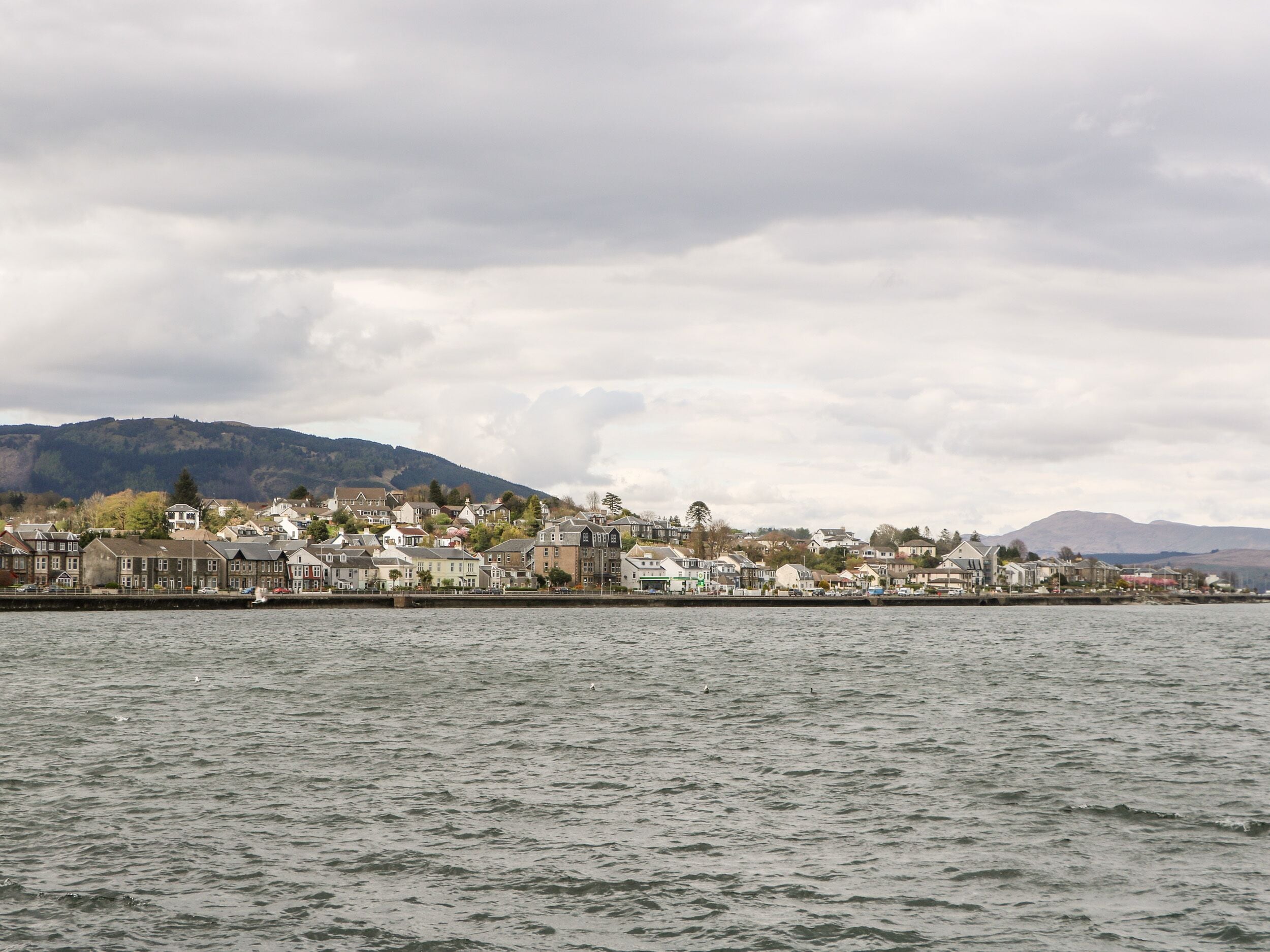 A coastal town with houses along the waterfront, a harbor, and hills in the background.