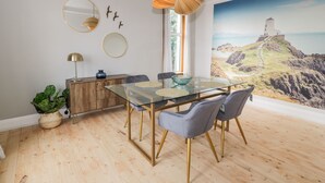 Un coin repas comprenant une table en verre, des chaises grises, un meuble en bois et un tableau représentant un paysage côtier.
