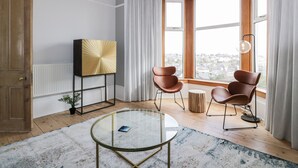 Un salon moderne avec une table basse en verre, deux fauteuils en cuir, un meuble décoratif doré et une grande fenêtre donnant sur un paysage urbain.