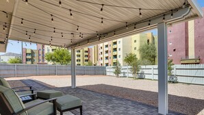 Ein überdachter Außenbereich mit einer Terrassentisch und Stühlen, einer Pergola mit Lichterketten und Blick auf mehrstöckige Gebäude im Hintergrund.