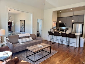 A modern living room with a sofa, coffee table, and bar stools. A kitchen with a refrigerator and microwave is visible in the background.