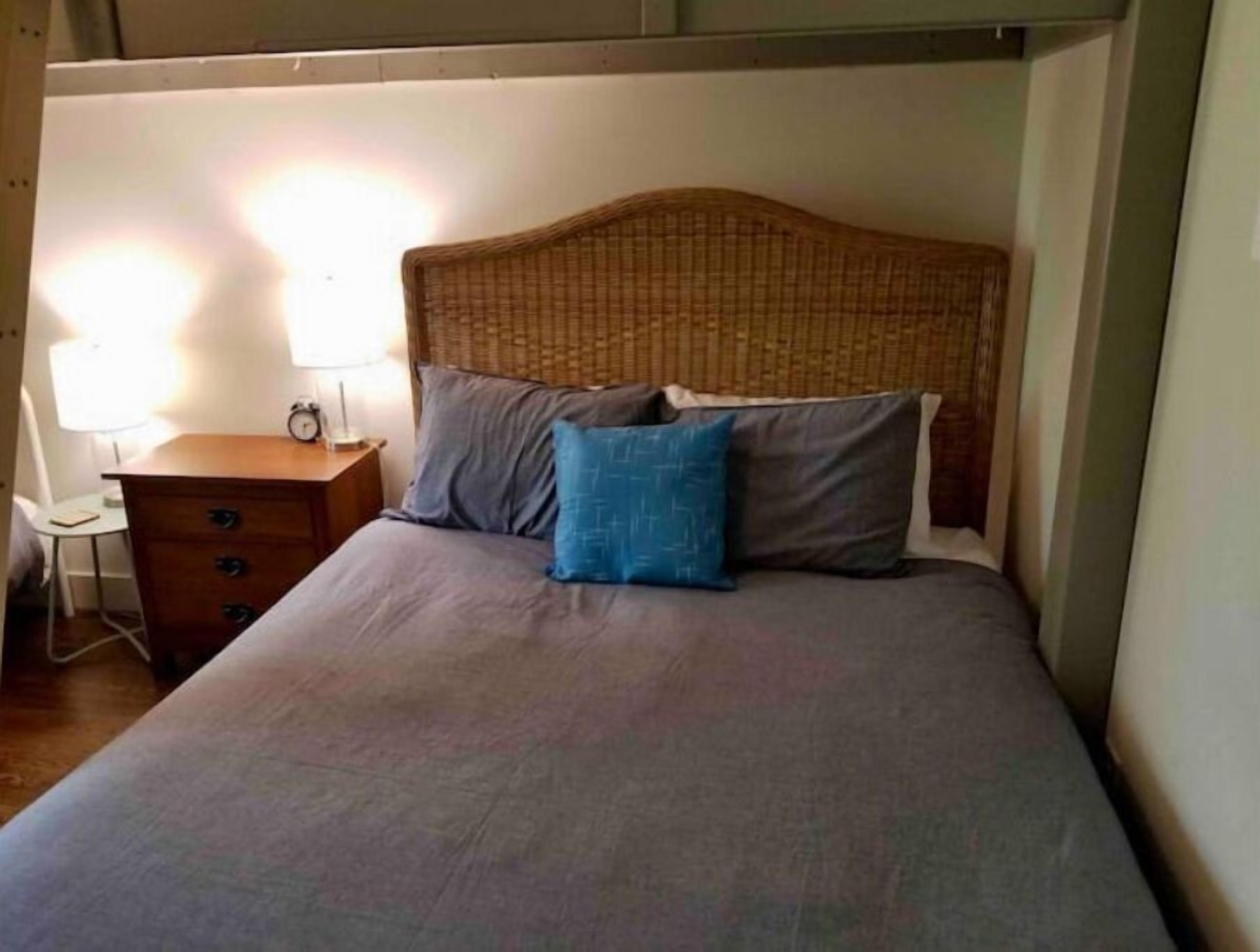 A bed with a woven headboard, a bedside table with a lamp, and a small desk with a chair.