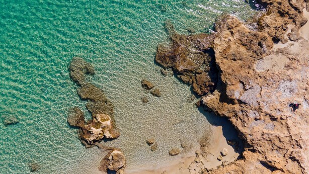Vista aérea de uma costa rochosa com águas turquesas cristalinas e áreas de praia arenosa.