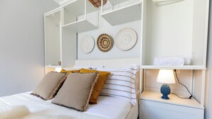 Cama bem arrumada com edredom branco e duas almofadas, abajur e uma prateleira pequena com uma toalha.