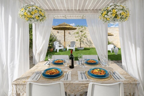 An outdoor dining area with white chairs, a table set with plates of pasta, wine glasses, and a bottle of wine.