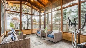 A covered outdoor area with wicker furniture, exercise equipment, and large windows offering views of trees and a body of water. - Swim Spa cottage on a Private Beach (Fort Erie)