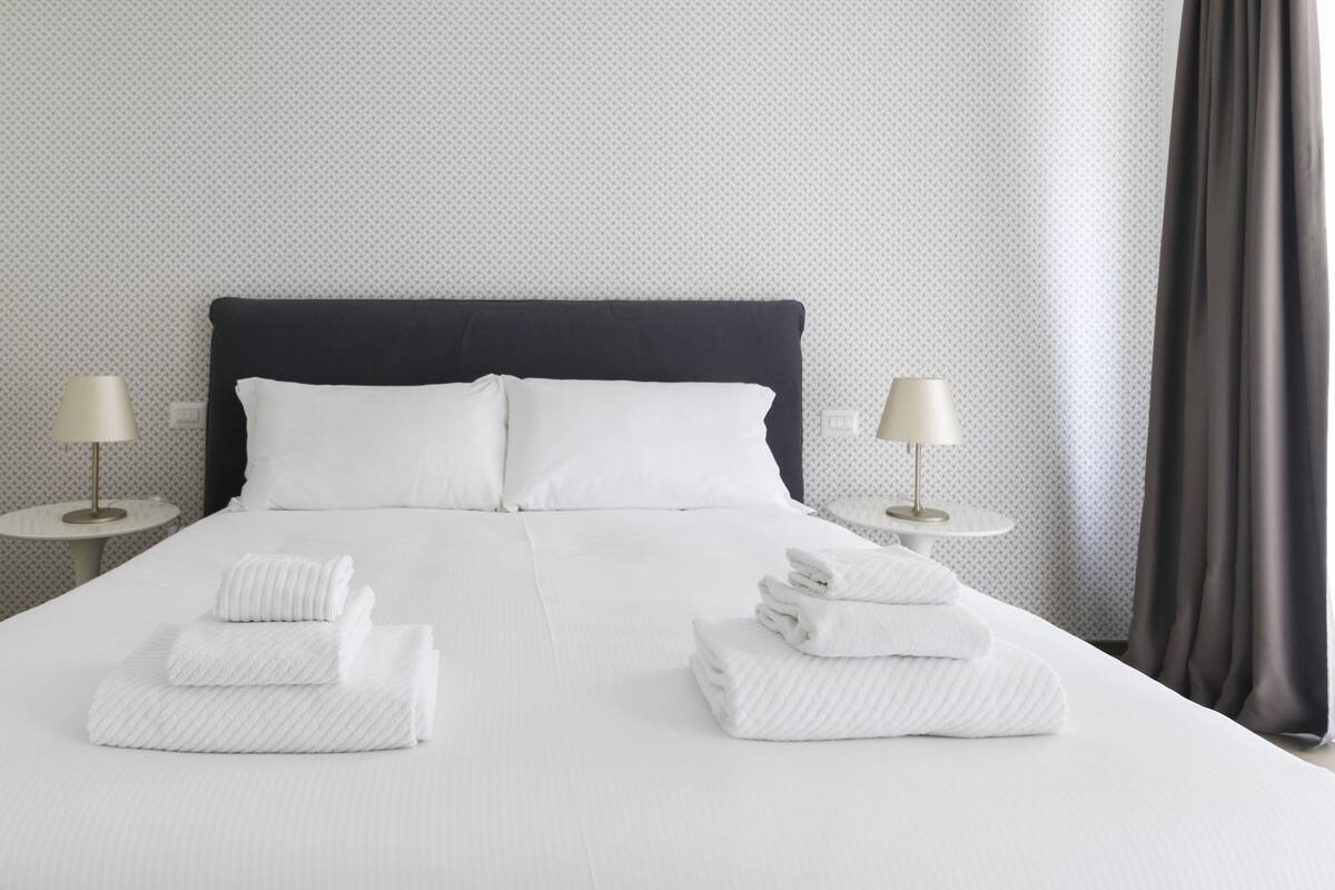 A neatly made bed with white linens and two towels folded at the foot of the bed. There are two bedside tables with lamps on either side of the bed.