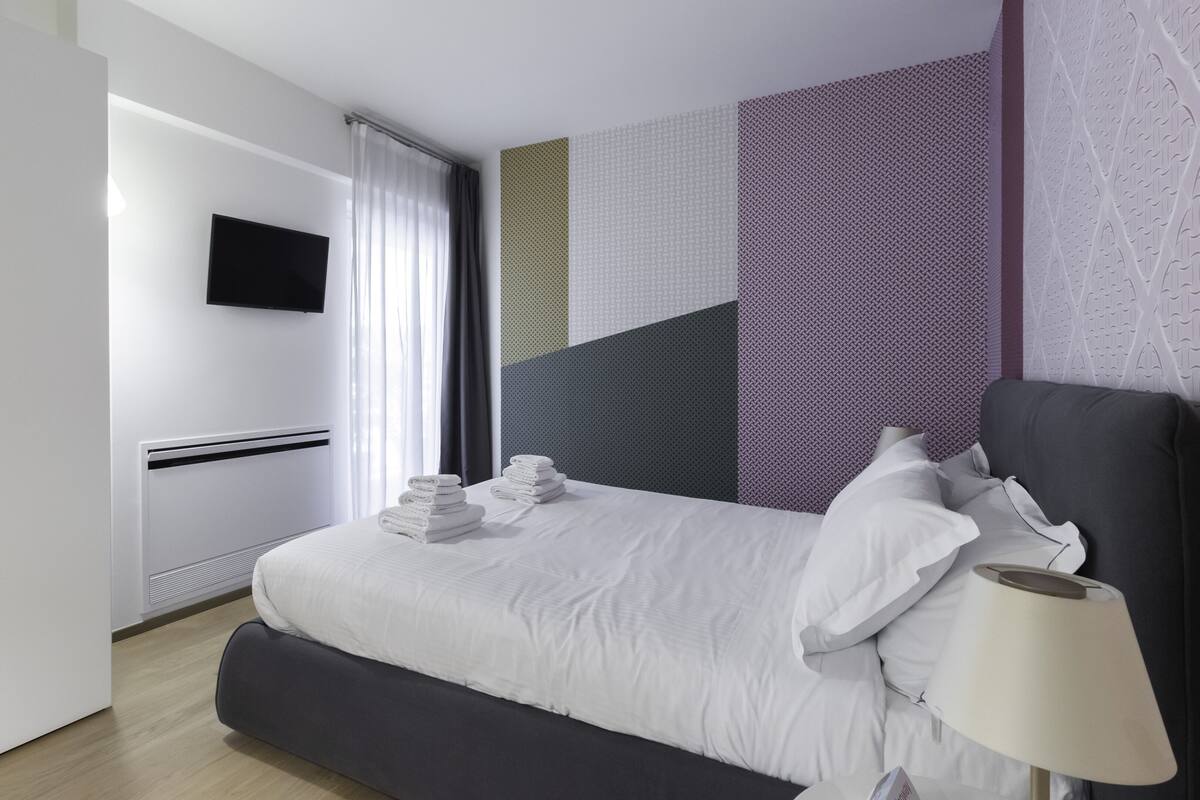 A modern hotel room with a large bed, a flat-screen TV mounted on the wall, and a neatly made bed with towels.