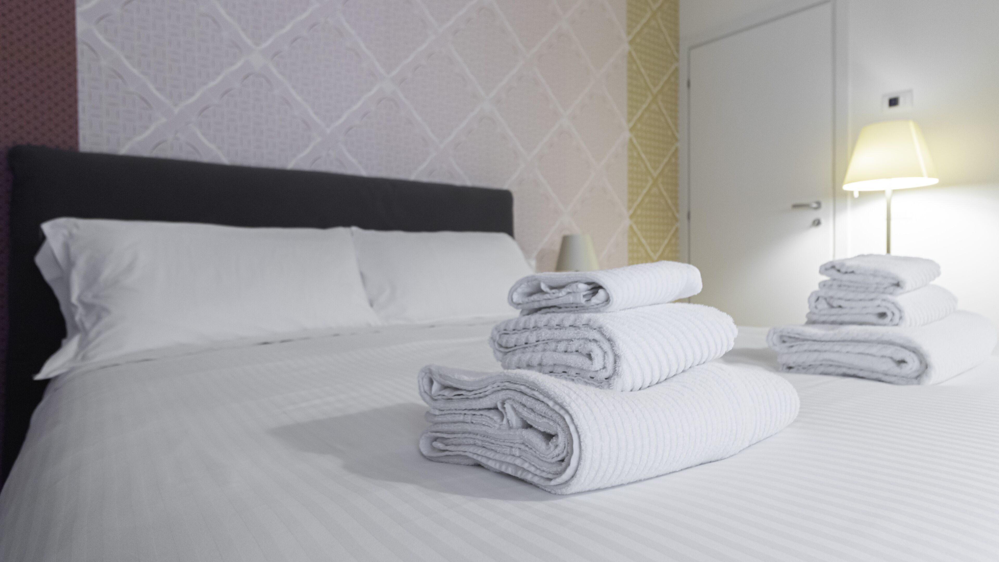 Una cama bien tendida con sábanas blancas, cabecera oscura y pared con diseño geométrico.