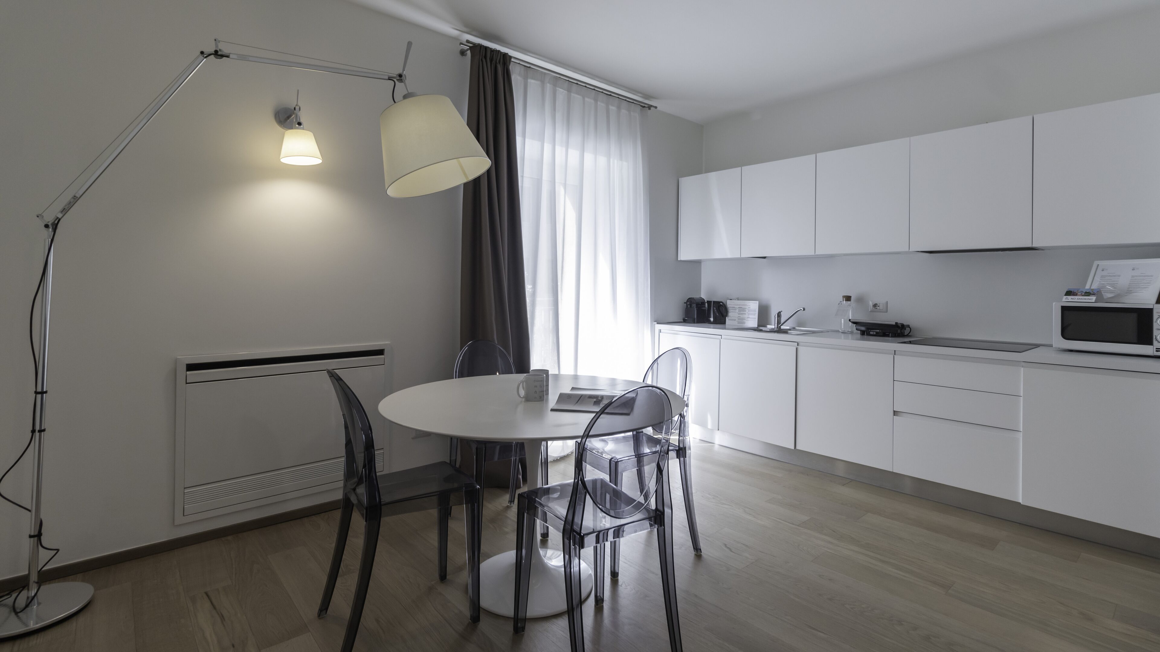 A modern kitchen with white cabinets, a round dining table, and two chairs. There is a floor lamp, a window with curtains, and a microwave on the counter.
