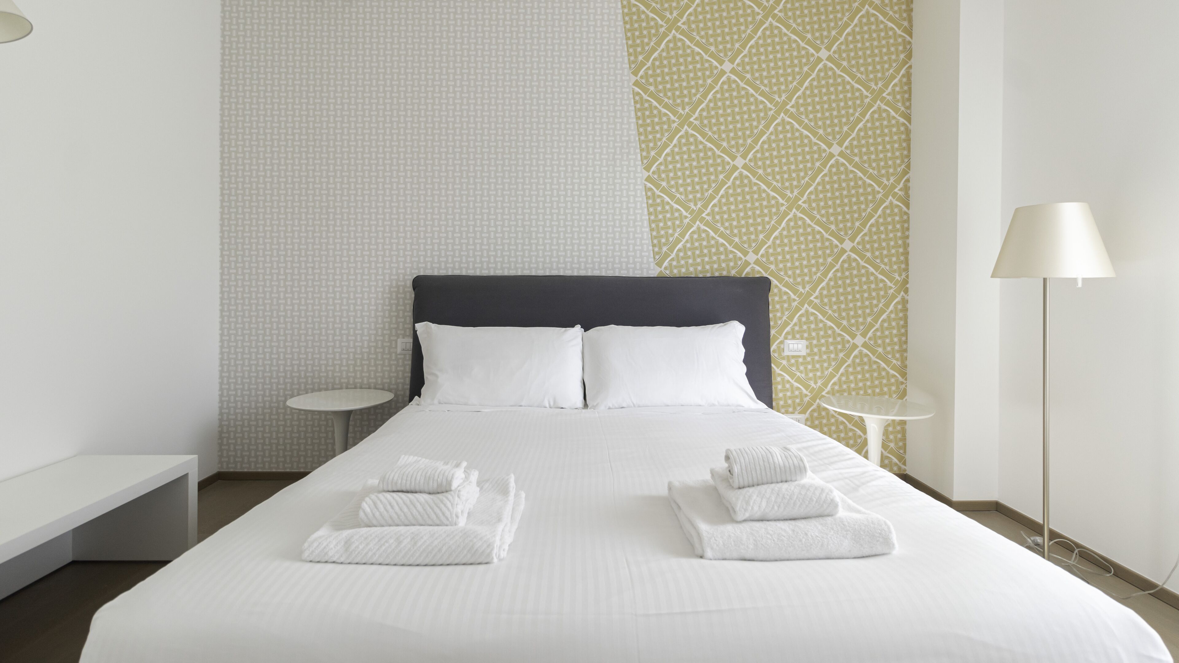 Un lit bien fait, avec du linge de lit et des serviettes blanches, une tête de lit sombre et un mur orné d’un motif.