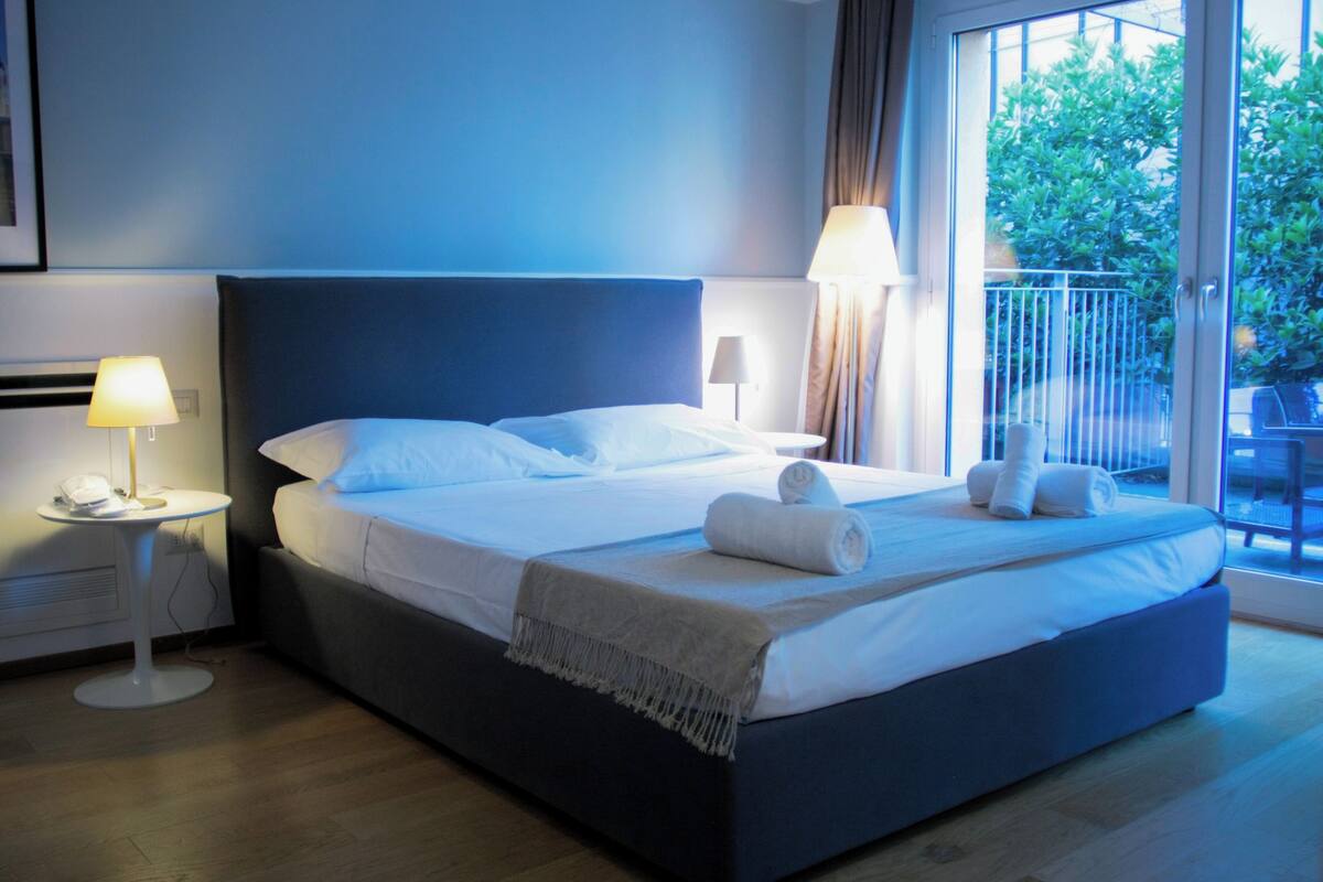 A neatly made bed with white linens and two rolled towels, a bedside table with a lamp, and a view of greenery through a large window.