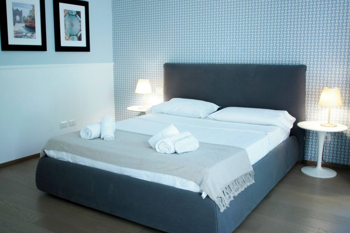 A neatly made bed with white linens and a grey headboard, two bedside lamps, and a wall with a patterned wallpaper.