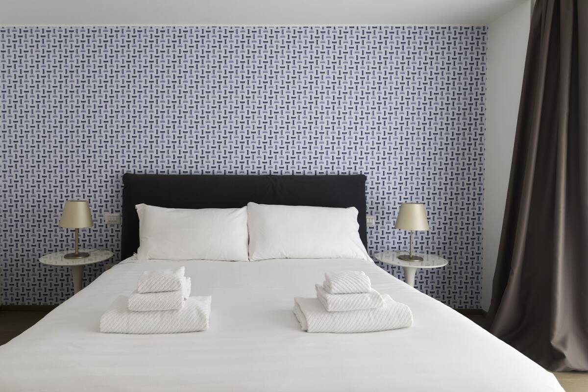 A neatly made bed with white linens, two bedside lamps, and a patterned wallpaper.