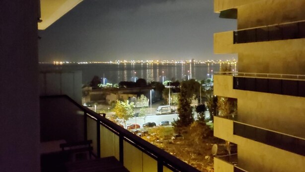Une vue nocturne depuis un balcon donnant sur une ville illuminée, avec des bâtiments éclairés qui se reflètent sur une étendue d’eau.