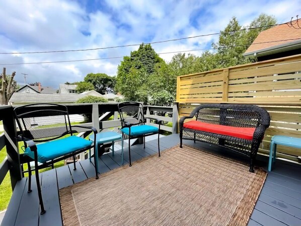 An outdoor deck with a wooden fence, a woven bench, and two chairs with cushions. - West Side Historic Home (Grand Rapids)