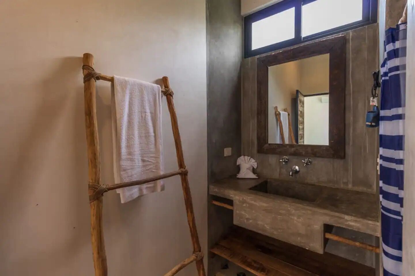 Un baño con una escalera de madera sosteniendo una toalla, un espejo grande sobre un lavabo y una cortina de ducha a rayas.