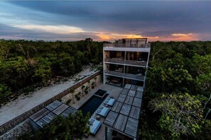 Un edificio moderno de varios pisos con piscina y zona de estar al aire libre, rodeado de abundante vegetación.