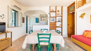 A dining area with a table, chairs, and a television. There is a sofa with a red cushion and a wooden shelf with decorative items. - Stone Hill House by BarbarHouse (Torre Vado)