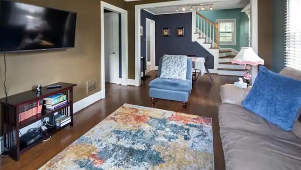 Una sala de estar con televisor de pantalla plana, estantería, alfombra estampada, sillón azul y una escalera que sube a otro nivel.