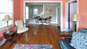 A living room with a wooden floor, a dining area with a table and chairs, a staircase, and a view of the outside. - Colorful Downtown Historic 2BR Shingle Home (Spartanburg)