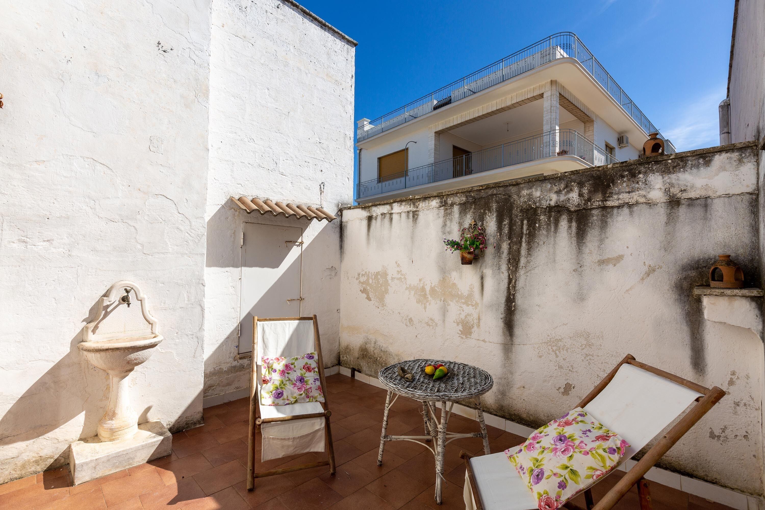 Eine kleine Außenpatio mit einer weißen Mauer, einer Steinfountain, einem Korbstuhl-Tisch mit Stühlen und einem Balkon darüber.