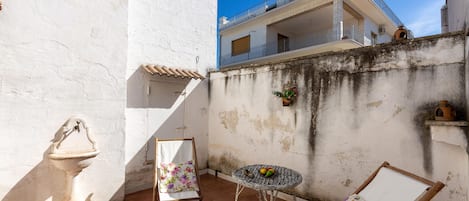 Eine kleine Außenpatio mit einer weißen Mauer, einer Steinfountain, einem Korbstuhl-Tisch mit Stühlen und einem Balkon darüber.
