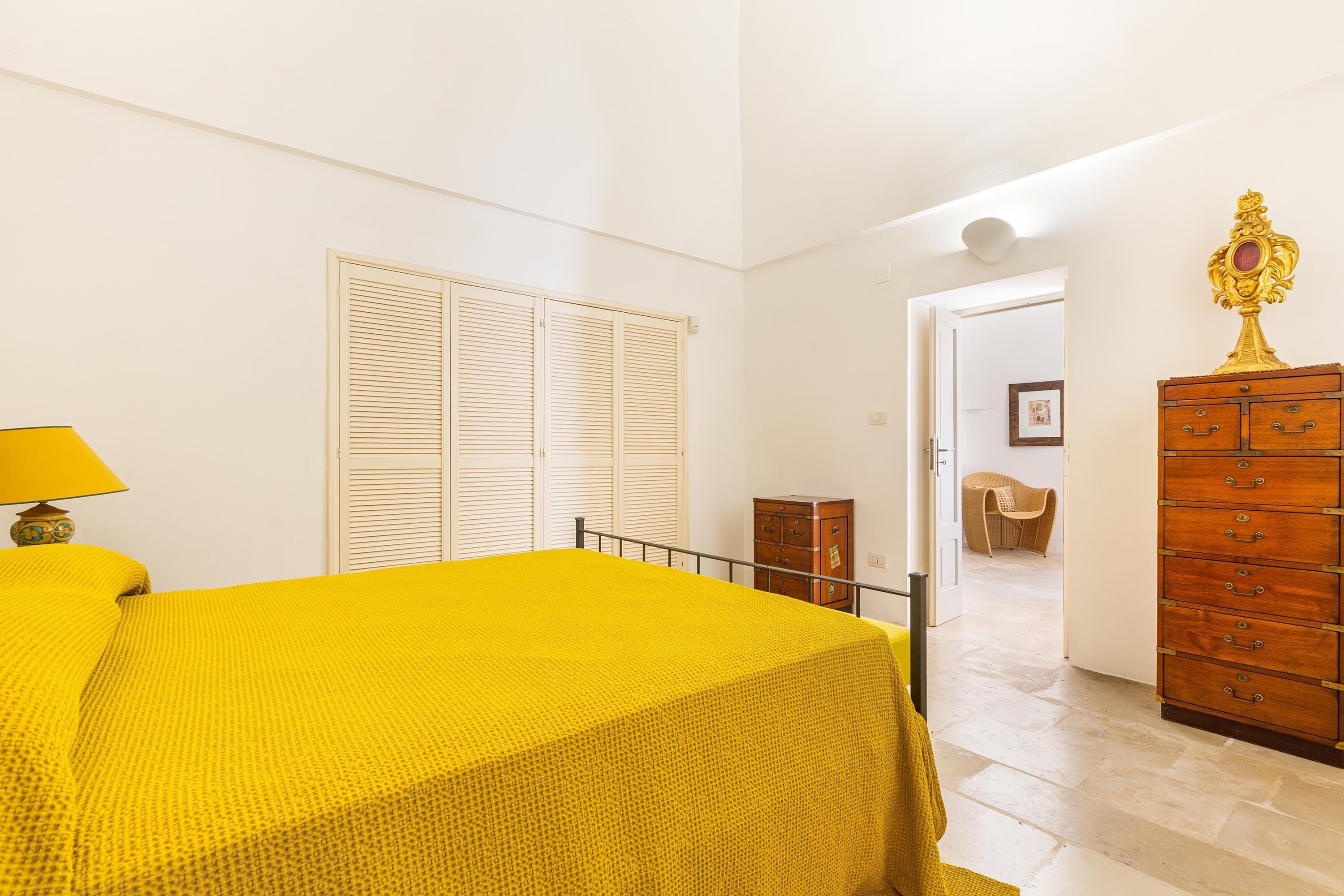 A room with a bed, a wooden dresser, a lamp, and a door leading to another room.