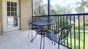 A balcony with a table and chairs, a door, and a view of a garden with palm trees. - Pretty & Welcoming Resort Condo (Kissimmee)