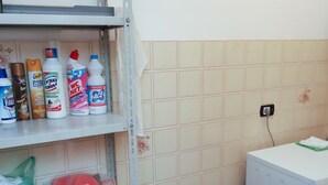 Une petite buanderie avec des étagères où sont rangés les produits de nettoyage, un lave-linge et une poubelle.
