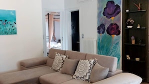 Een woonkamer met een beige bank, gekleurde kussens, een schilderij van bloemen en een boekenkast met decoratieve objecten.