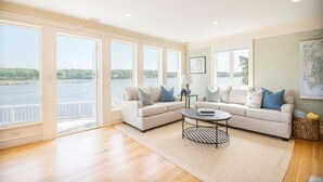 Ein geräumiges Wohnzimmer mit großem Fenster und Blick auf ein Gewässer, ein beiges Sofa, ein Couchtisch und eine Karte an der Wand.