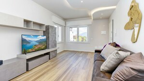 Ein modernes Wohnzimmer mit Flachbildfernseher, eingebauten Regalen, einem grauen Sofa und einem großen Fenster.