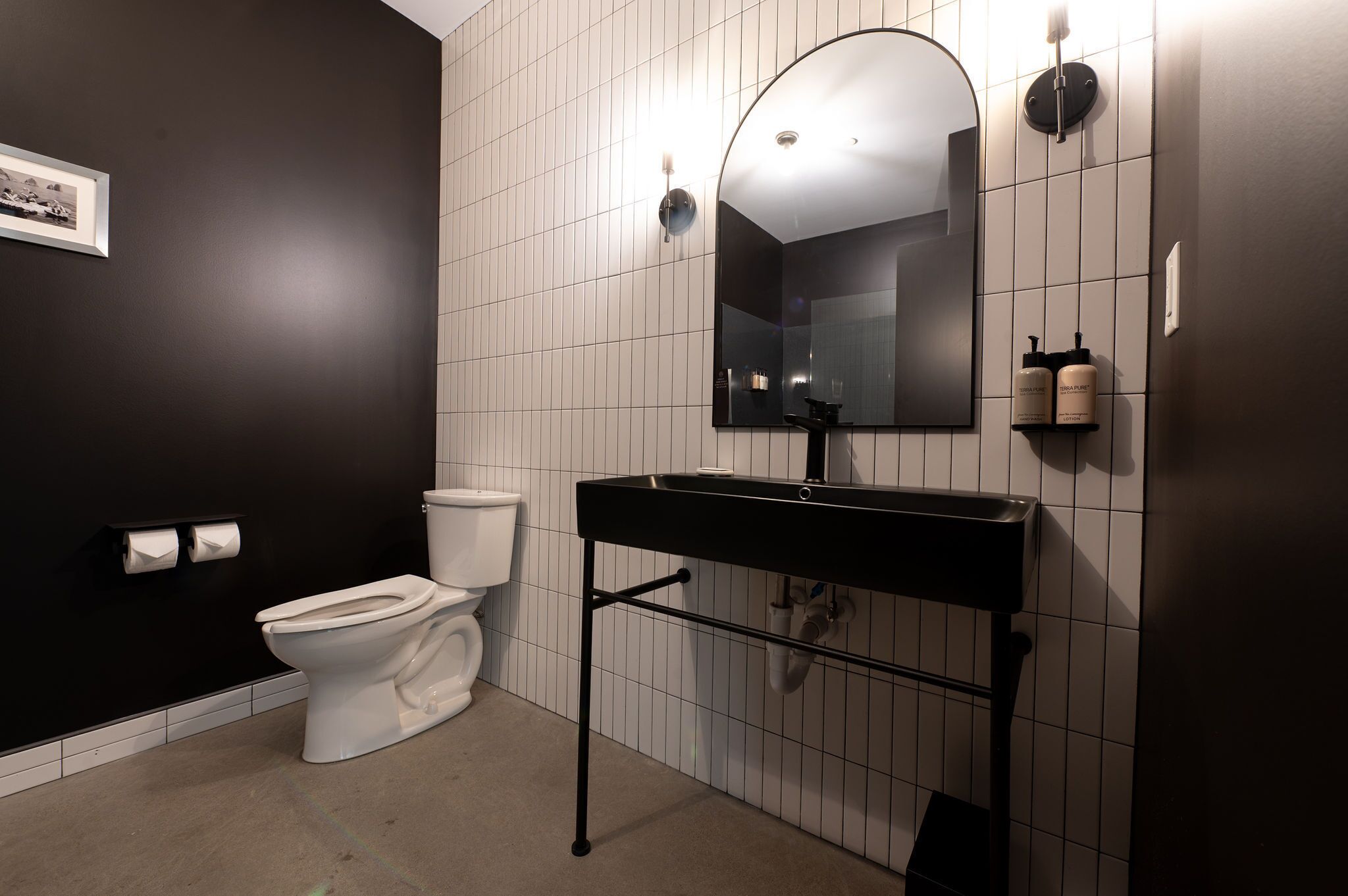 A modern bathroom with a black sink, white tiles, a toilet, and a framed picture on the wall.