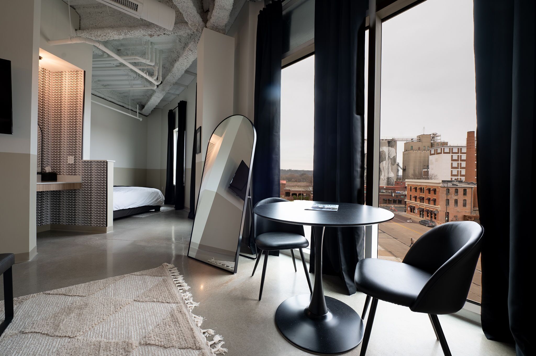 A modern hotel room with a bed, a round table with chairs, a large window with curtains, and a city view.