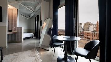 A modern hotel room with a bed, a round table with chairs, a large window with curtains, and a city view.