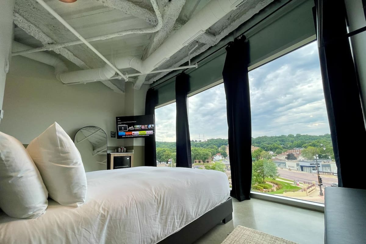 A modern hotel room with a large bed, a flat-screen TV, and a view of a cityscape through large windows.